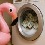ufblasbarer pinker Flamingo blickt neugierig in eine Waschmaschine, die mit Geldscheinen und schäumendem Wasser gefüllt ist – humorvolle Darstellung des Themas Geldwäsche.